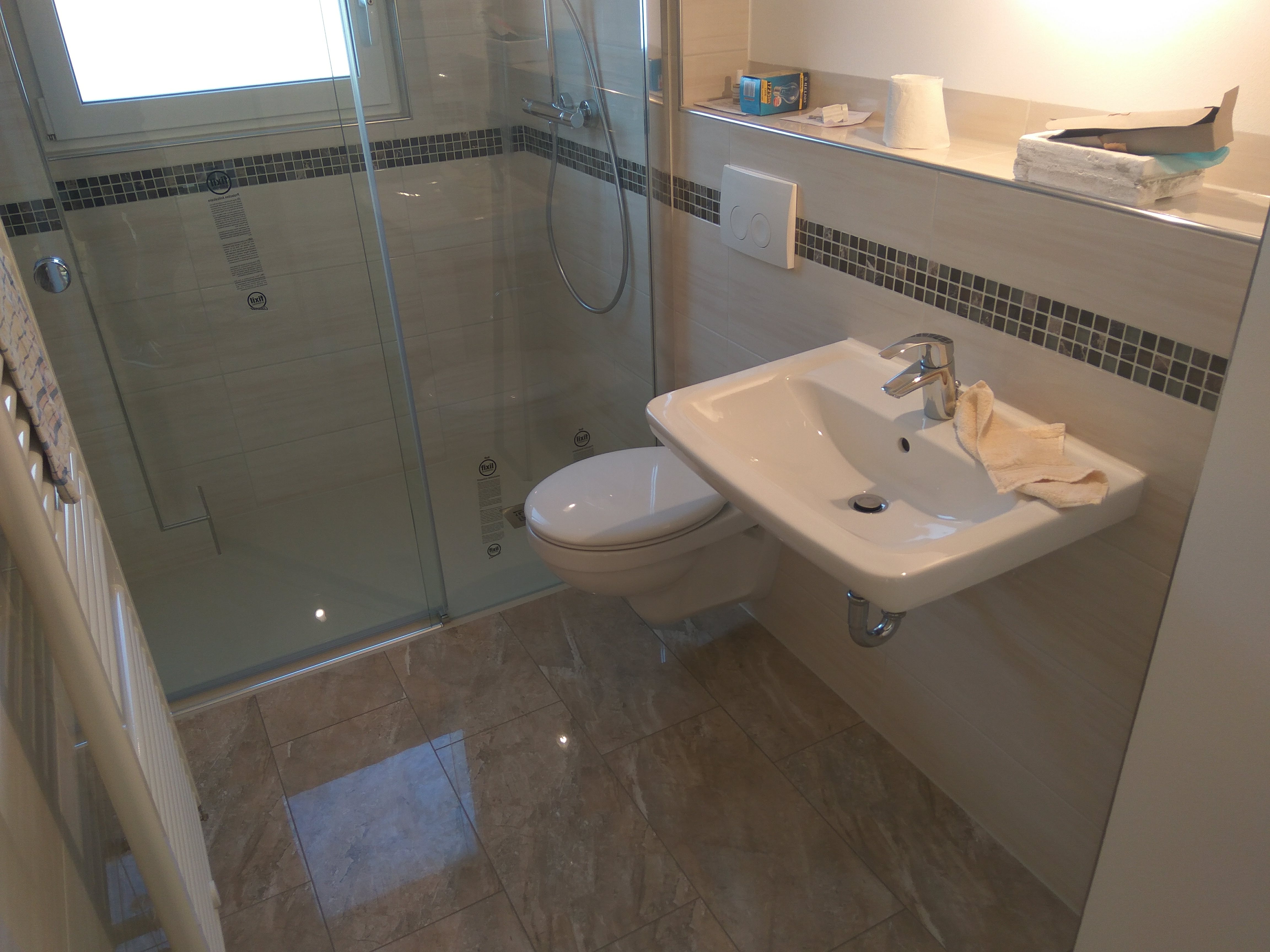 Nowoczesna łazienka z kabiną prysznicową ze szklanymi drzwiami, wiszącą toaletą i umywalką z baterią, wykończona beżowymi płytkami z dekoracyjnym pasem mozaiki.