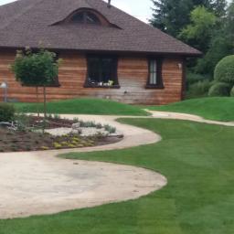 Tom-Kar - Kręta ścieżka z jasnych płyt prowadząca przez trawnik obok drewnianego domu z brązowym dachem i rabatą z kamieniami i niskimi roślinami.