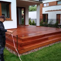 Montaż drewnianego tarasu z egzotycznego drewna przy nowoczesnym domu, pracownik ustawia poziom, widoczne schody tarasowe i fragment ogrodu.