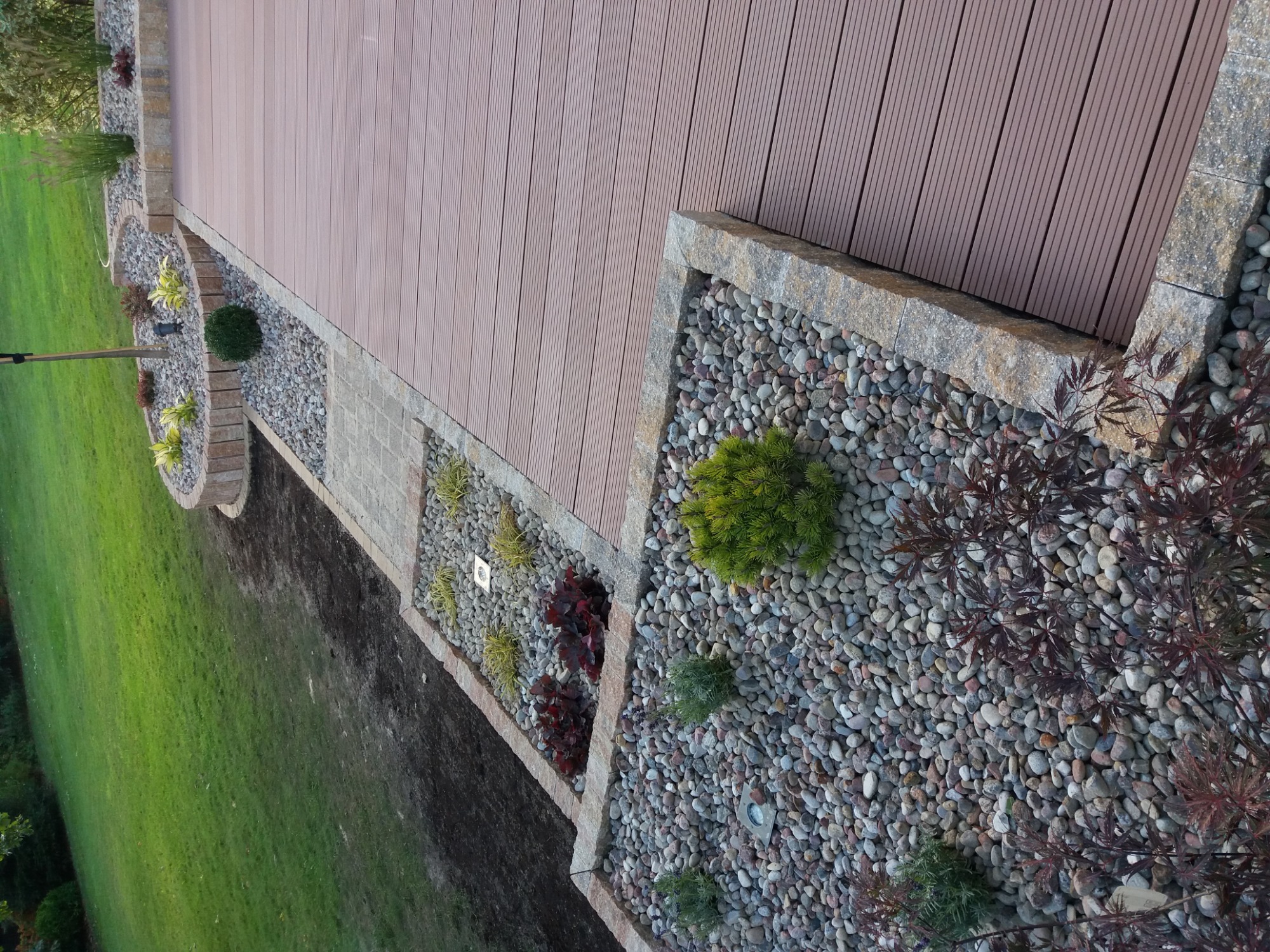 Kompozytowy taras w kolorze drewna obramowany kamienną rabatą wypełnioną drobnymi kamieniami i ozdobnymi roślinami, widoczne punkty oświetleniowe w rabacie.