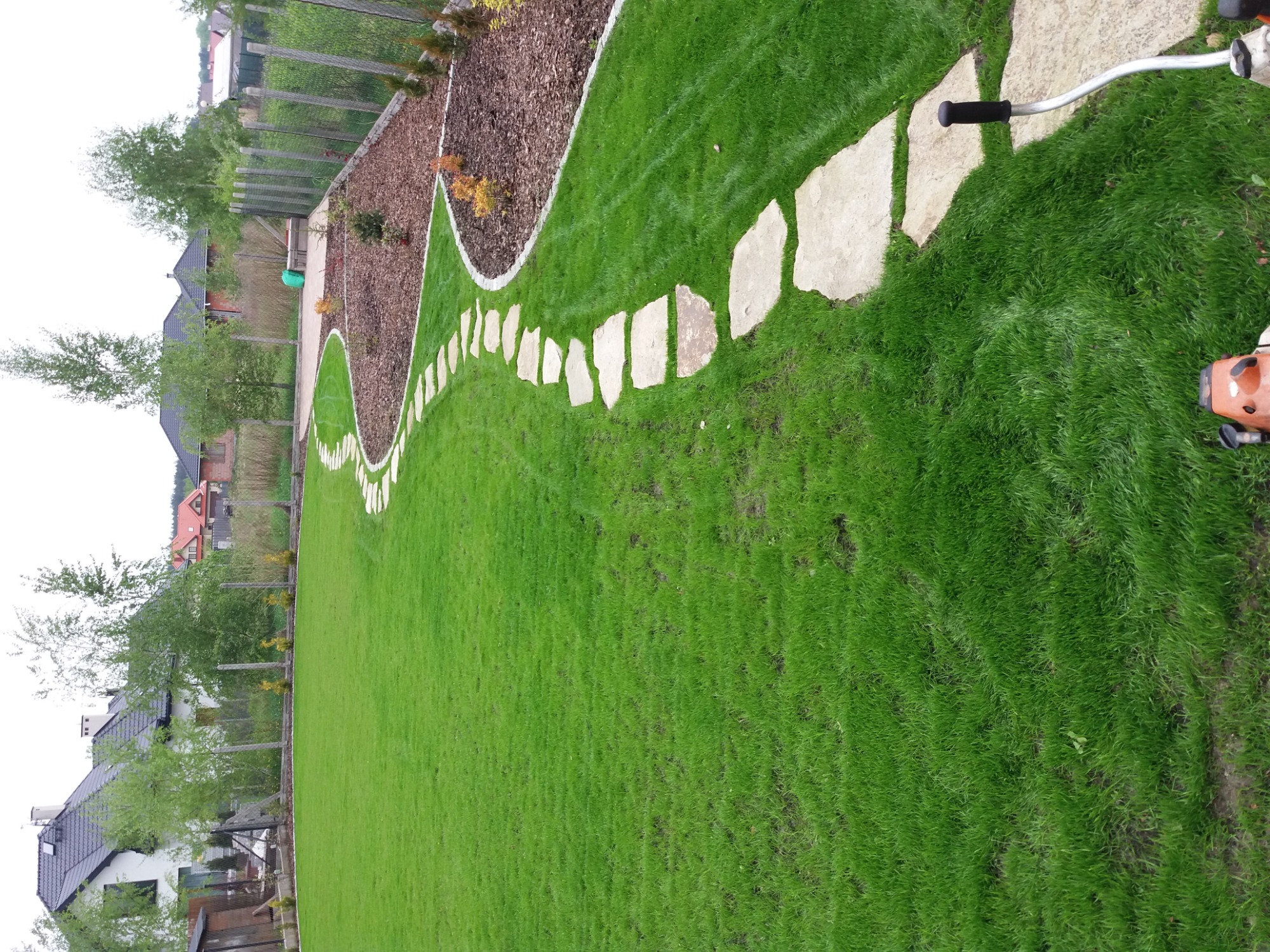 Kaskadowa ścieżka z nieregularnych płyt kamiennych na trawniku, obok rabaty z korą i obrzeżem, widok z góry z fragmentem kosiarki.