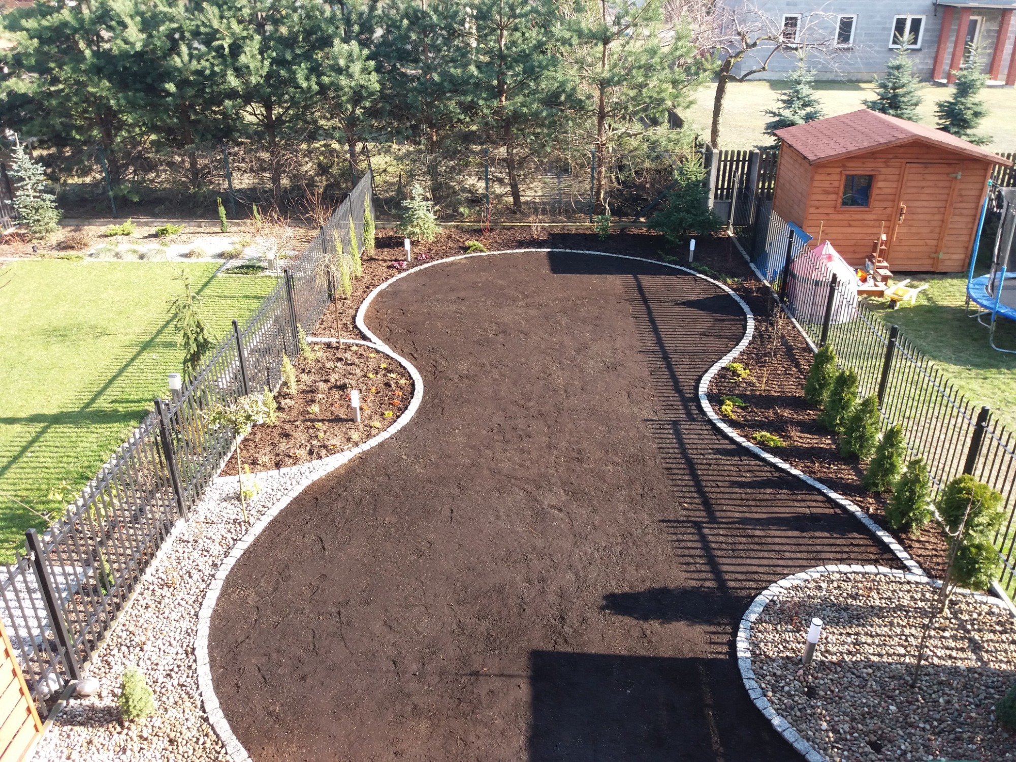 Przygotowany teren pod trawnik lub nasadzenia, obrzeża z jasnej kostki granitowej, widok z góry na posesję z metalowym ogrodzeniem i drewnianą altaną.