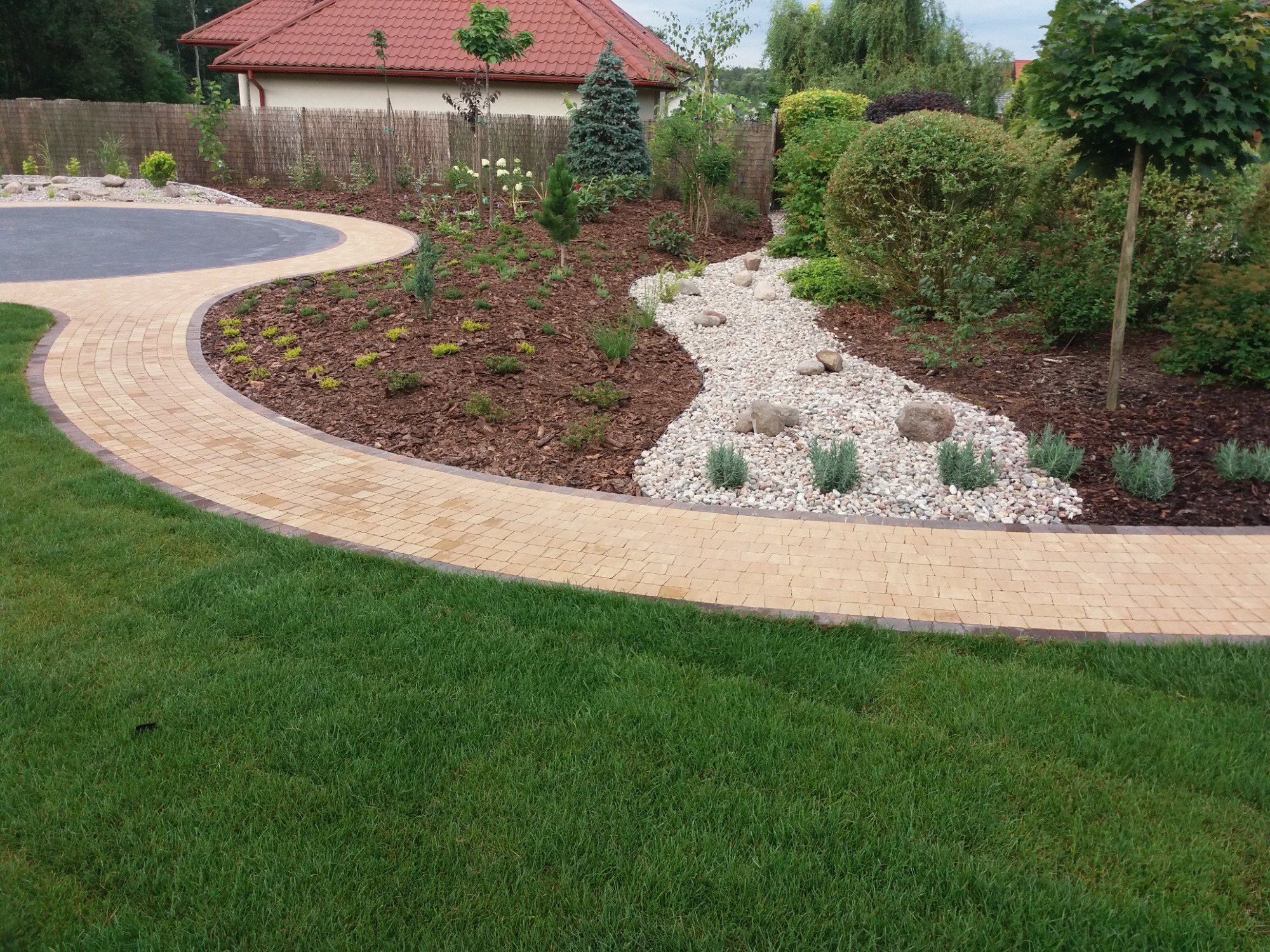 Zakrzywiony chodnik z jasnej kostki brukowej z ciemniejszą obwódką, prowadzący przez zadbany ogród z korą, kamieniami i różnorodną roślinnością.