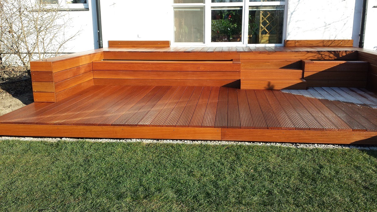 Nowoczesny taras z drewna kompozytowego, zintegrowany z budynkiem, otoczony trawnikiem i drobnym żwirem.