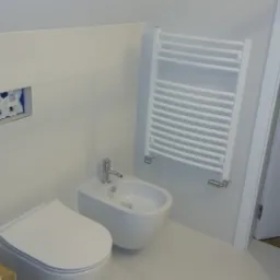 Nowoczesna, minimalistyczna łazienka z białymi płytkami, wiszącą toaletą i bidetem, grzejnikiem drabinkowym oraz widoczną instalacją podtynkową w ścianie.