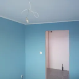 Pomieszczenie po malowaniu ścian na niebiesko z białym wykończeniem sufitu, widoczne przewody od lampy sufitowej, szara podłoga i przejście do kolejnego pomieszczenia.