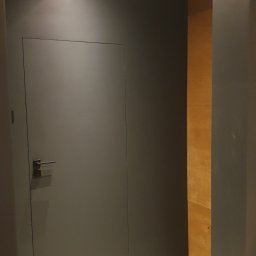 Drzwi w kolorze ścian bez futryn, a na ścianie bejcowana sklejka