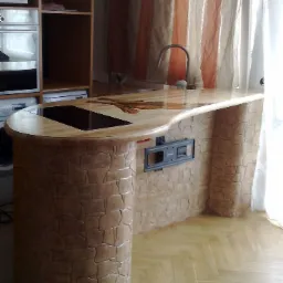 Wyspa kuchenna z blatem z jasnego drewna, wbudowaną płytą indukcyjną i dekoracyjną okładziną imitującą kamień na podstawie, obok zlewozmywak i okno z zasłonami.