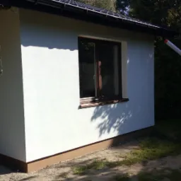 Świeżo pomalowana, biała elewacja domu z brązowymi akcentami przy oknach i podstawie, widoczny fragment rynny i drzwi wejściowe, otoczenie z zielenią.