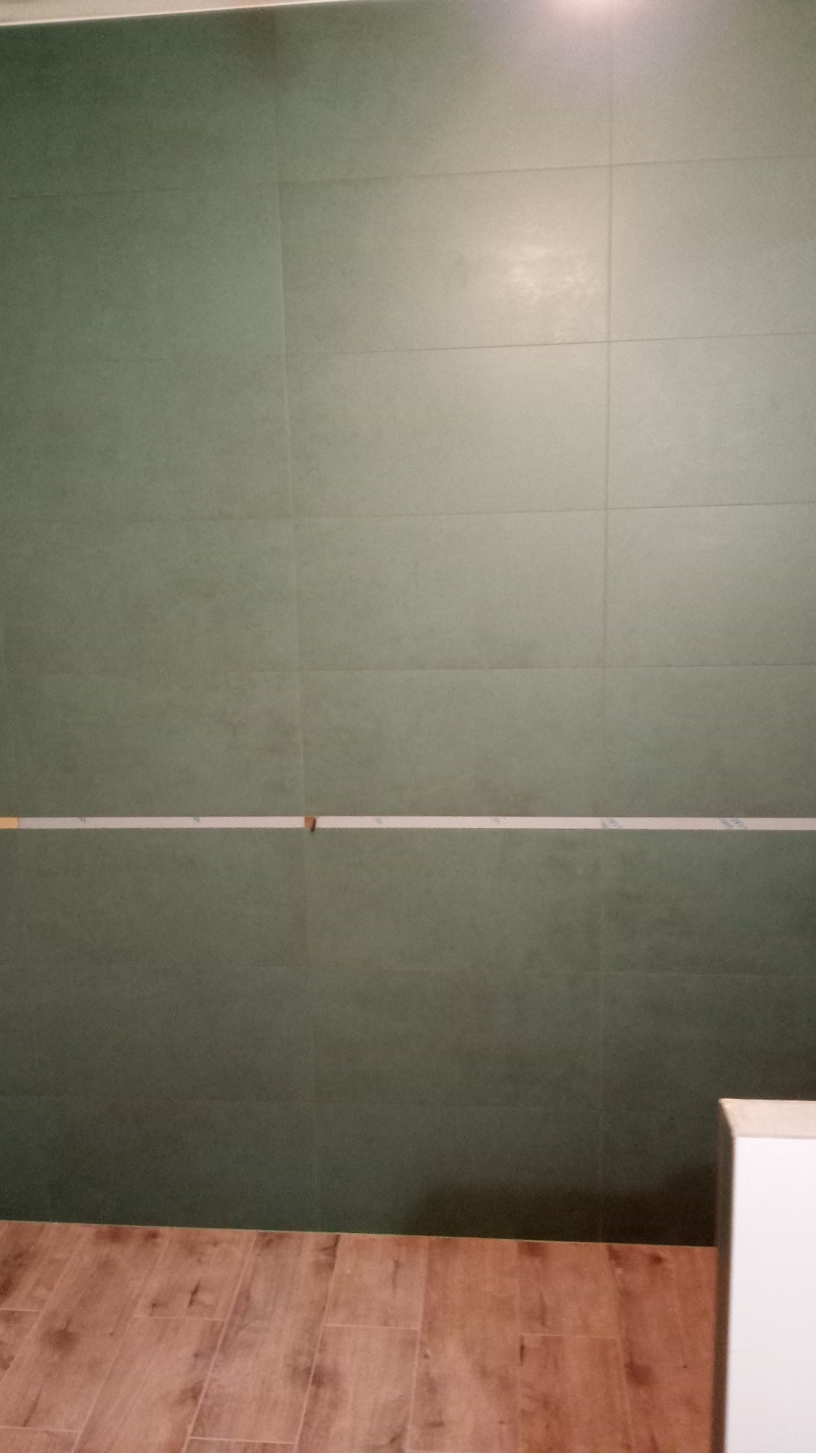 Ściana wyłożona dużymi, matowymi płytkami w odcieniach zieleni, z poziomym pasem przygotowanym pod fugowanie, oraz podłoga wyłożona panelami imitującymi drewno.