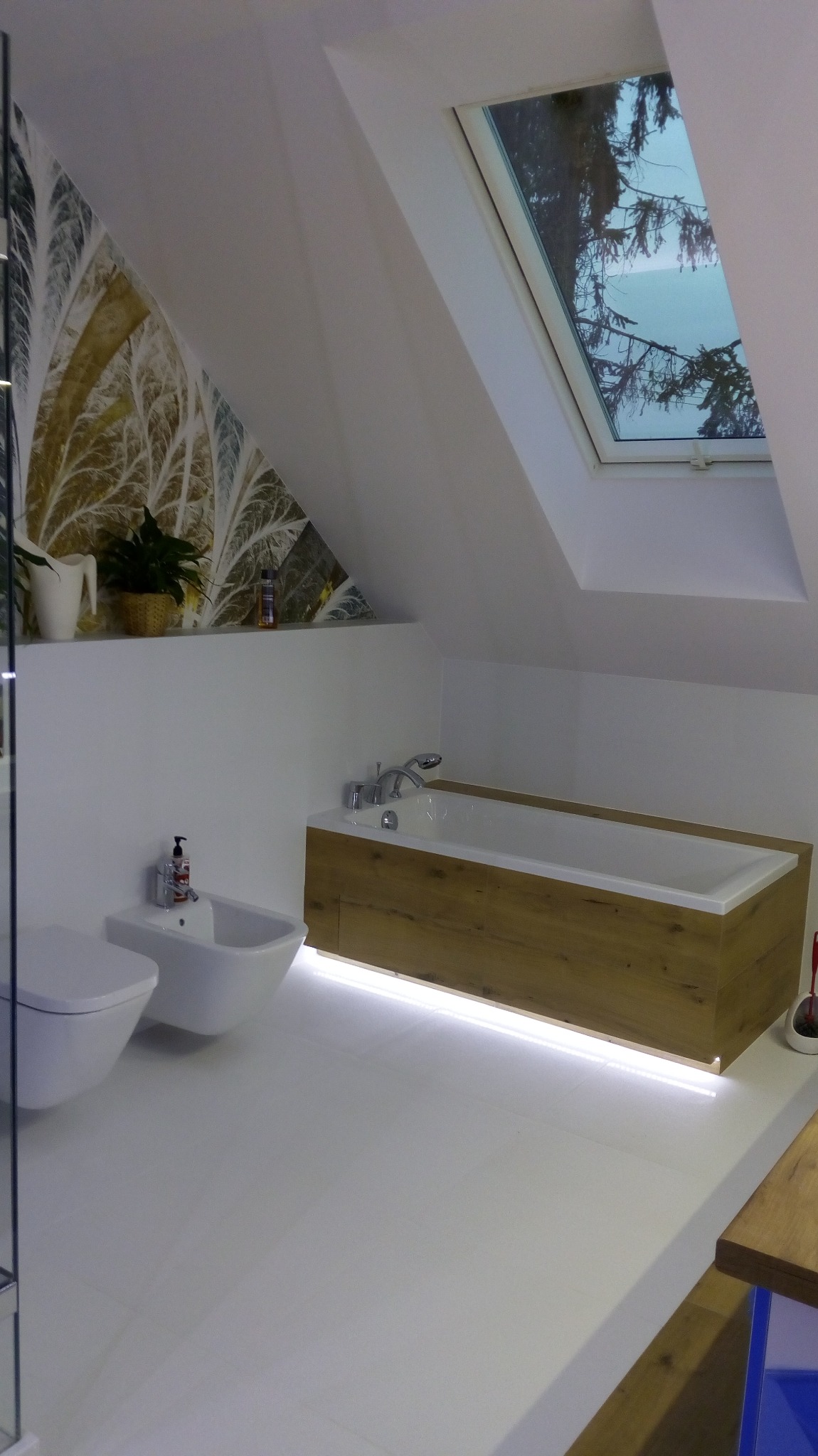 Nowoczesna łazienka na poddaszu z białą armaturą, drewnianą obudową wanny z podświetleniem LED, wzorzystą tapetą i oknem dachowym.