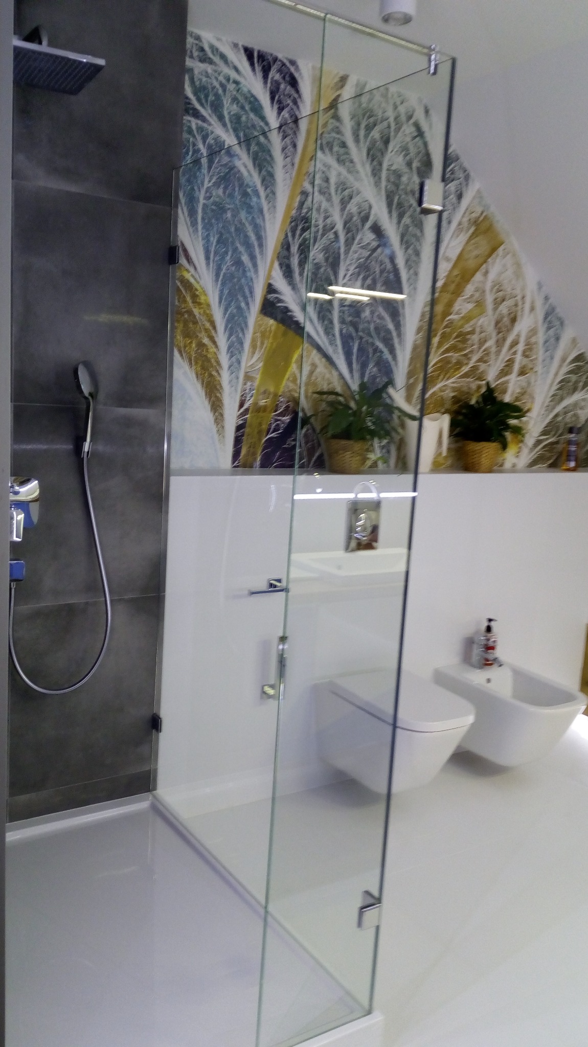 Nowoczesna łazienka z szarą strefą prysznicową z deszczownicą i szklaną kabiną, białą ceramiką sanitarną (toaleta, bidet) oraz ścianą z dekoracyjną tapetą w motywy drzew.