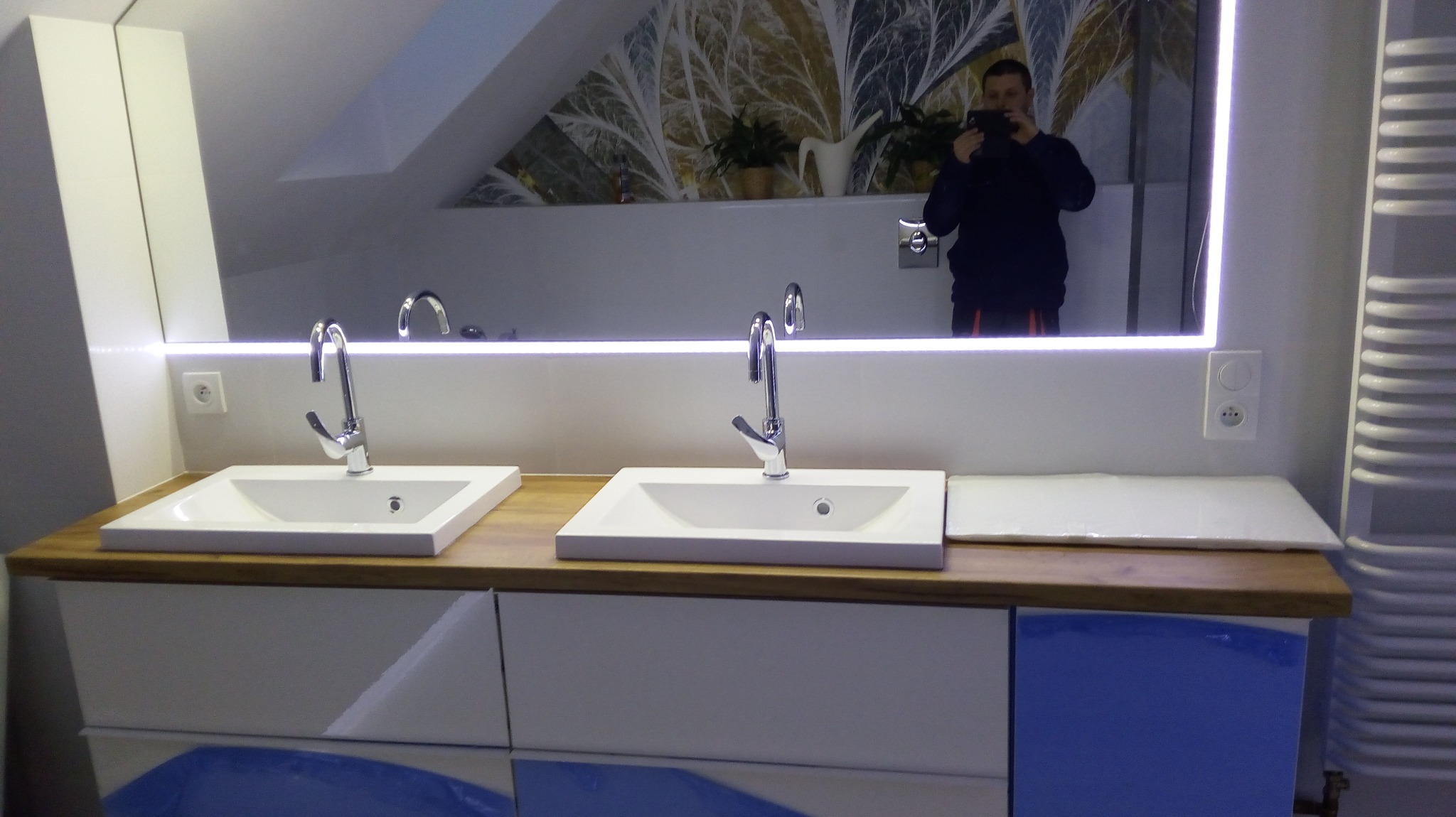 Nowoczesna łazienka z dwoma umywalkami nablatowymi, drewnianym blatem i szafką w kolorze białym i niebieskim, oświetlone lustro z odbiciem osoby robiącej zdjęcie.