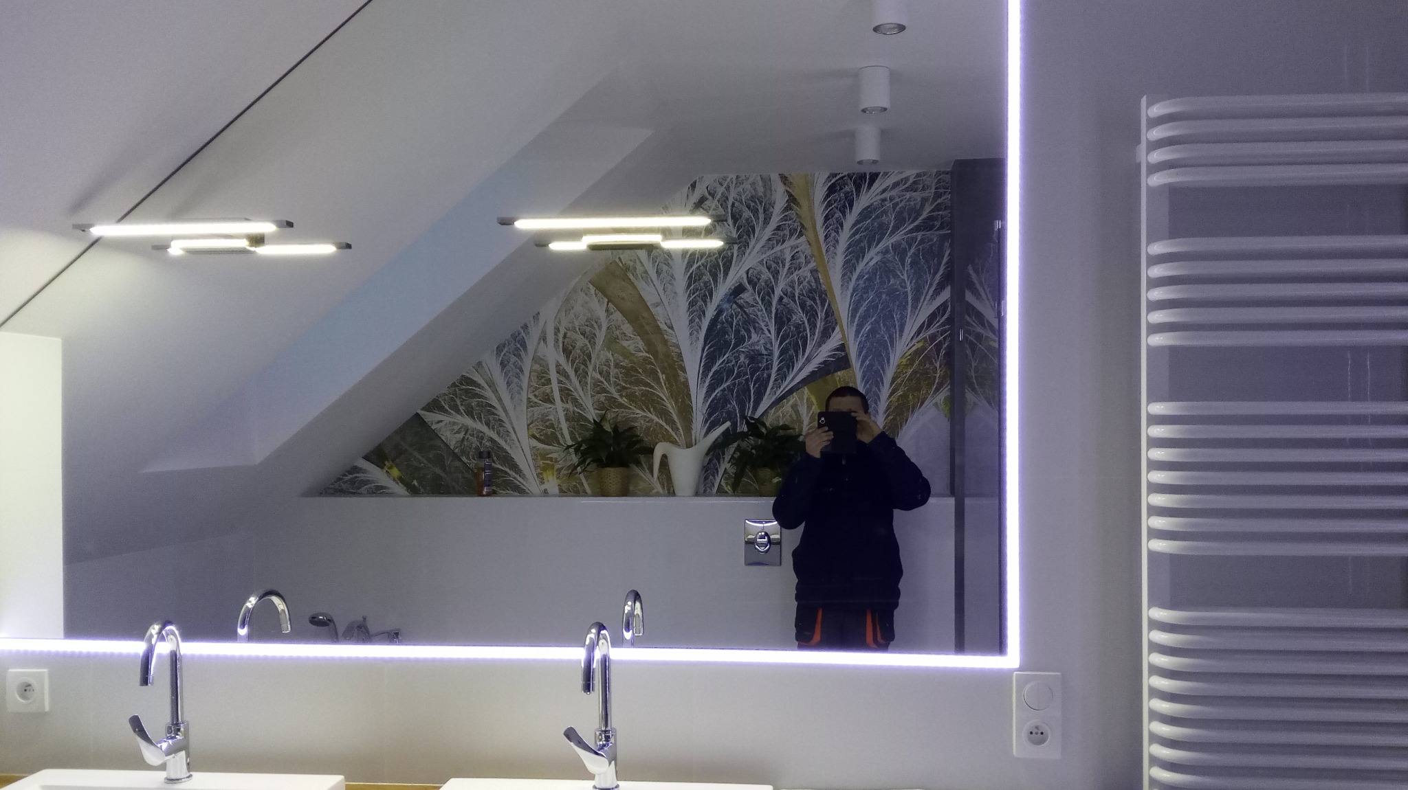 Nowoczesna łazienka z podświetlanym lustrem LED, wzorzystą tapetą z motywem roślinnym, grzejnikiem drabinkowym i dwiema umywalkami; osoba robi zdjęcie w odbiciu lustra.