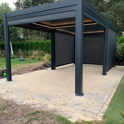 PPHU Rumin Marcin - Nowoczesna, grafitowa pergola metalowa z roletami przeciwsłonecznymi i oświetleniem LED, ustawiona na wybrukowanym tarasie w ogrodzie. Widoczny robot koszący w tle.