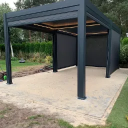 Nowoczesna, grafitowa pergola metalowa z roletami przeciwsłonecznymi i oświetleniem LED, ustawiona na wybrukowanym tarasie w ogrodzie. Widoczny robot koszący w tle.