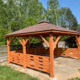 Drewniana altana z brązowym dachem na kostce brukowej, z częściowym ażurowym wypełnieniem ścianek, widok na zieleń i grill murowany w tle.