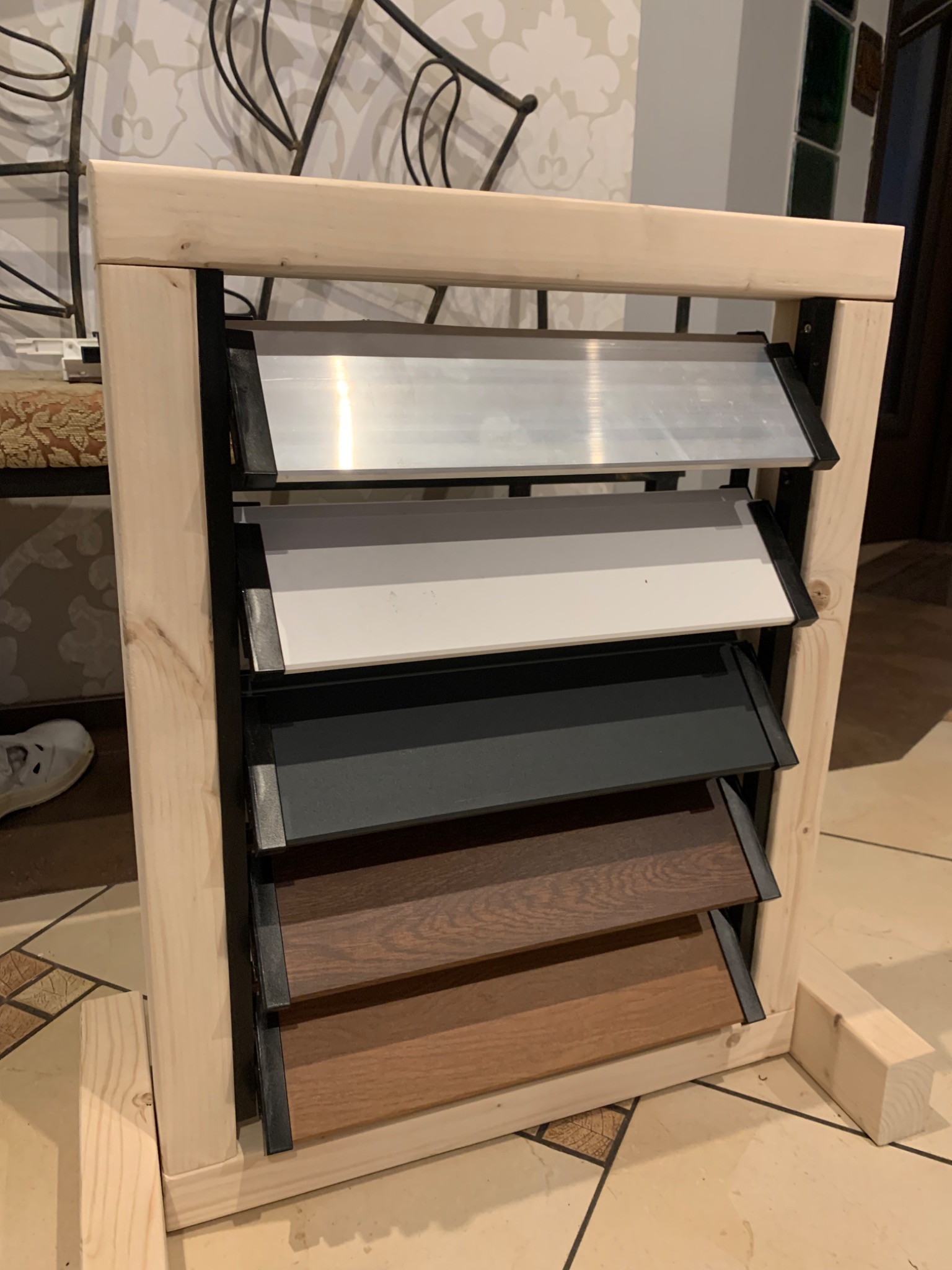 Ekspozytor z próbkami żaluzji zewnętrznych w różnych kolorach i materiałach, umieszczony w drewnianej ramie.