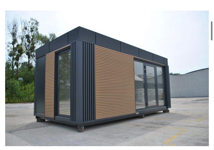 Nowoczesny, modułowy budynek z szarymi ramami okiennymi i panelami imitującymi drewno, posadowiony na betonowej powierzchni, z widocznym fragmentem zieleni w tle.