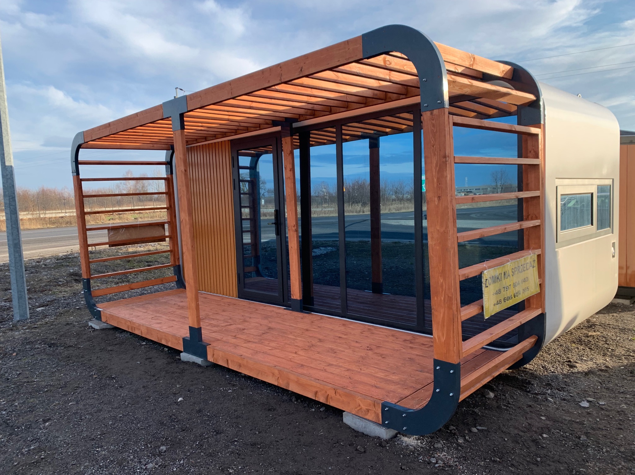 Nowoczesny modułowy budynek z elementami drewnianej pergoli i przeszklonymi drzwiami, umieszczony na betonowych fundamentach, z widoczną tabliczką 'Domki na sprzedaż'.