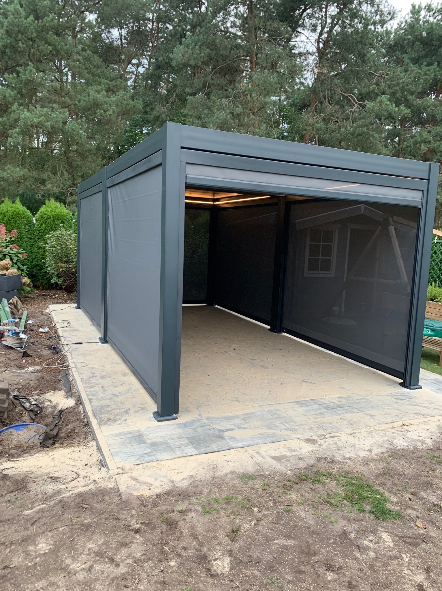 Nowoczesna, szara konstrukcja z metalu z roletami tekstylnymi, prawdopodobnie pergola, na wybrukowanym patio, otoczona zielenią, z widocznymi śladami prac budowlanych w tle.