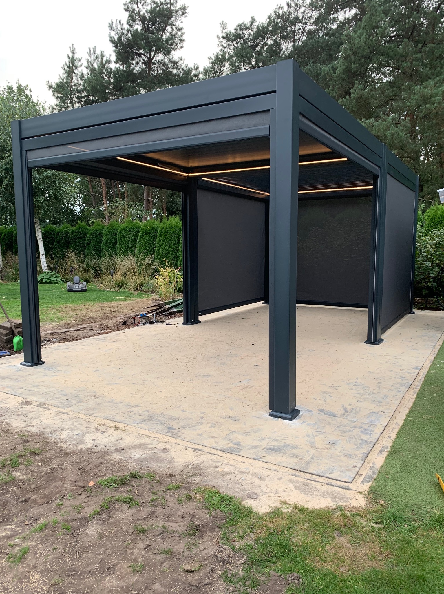 Nowoczesna, grafitowa pergola metalowa z roletami przeciwsłonecznymi i oświetleniem LED, ustawiona na wybrukowanym tarasie w ogrodzie. Widoczny robot koszący w tle.