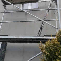 Szare rusztowanie na tle świeżo otynkowanej elewacji budynku, widoczne okna z siatką ochronną i fragment iglaka.