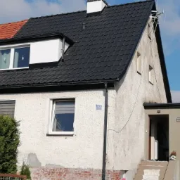 Dom z częściowo wymienionym dachem z czarnej dachówki, widoczna różnica kolorystyczna w porównaniu do starej, czerwonej części dachu. Elewacja w kolorze jasnego kremu, z widocznymi śladami zabrudzeń.