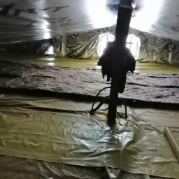 Poddasze z widoczną izolacją z wełny mineralnej, folią paroizolacyjną na podłodze i folią aluminiową na dachu, przez którą przechodzi czarna rura wentylacyjna.