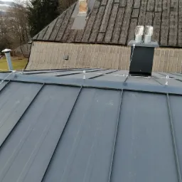 Nowy, grafitowy dach z blachy na tle starszego dachu z widocznym kominem i elementami zabezpieczeń przeciwoblodzeniowych.