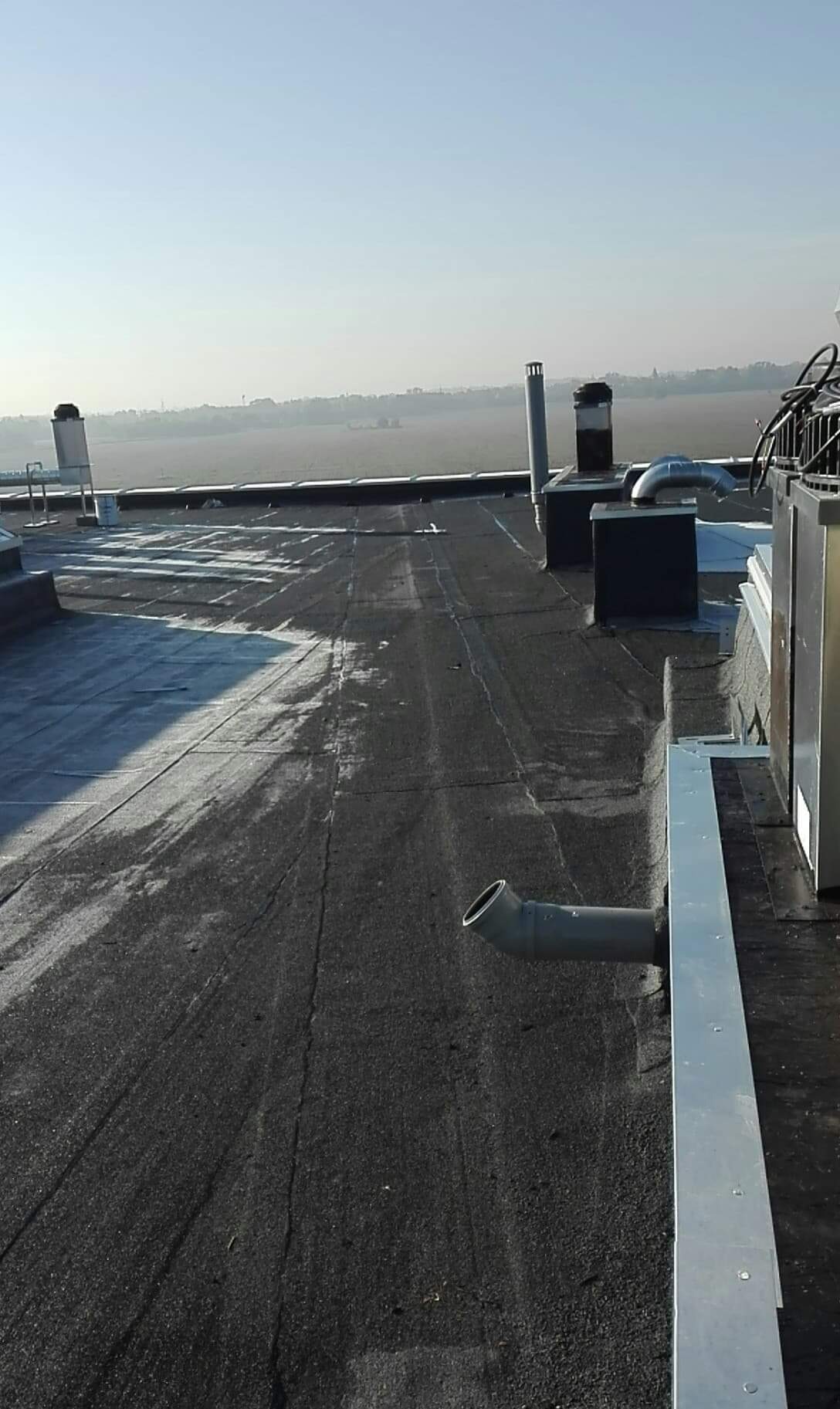 Płaski dach pokryty papą z widocznymi kominami wentylacyjnymi, obróbkami blacharskimi i fragmentami instalacji na tle zamglonego krajobrazu.