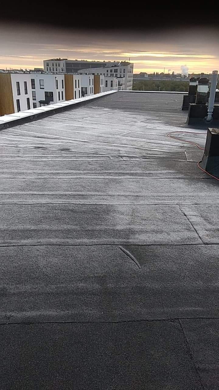 Widok z góry na płaski dach pokryty papą termozgrzewalną, z widocznymi łączeniami i niewielkimi uszkodzeniami, budynki mieszkalne w tle, pomarańczowy przewód na dachu