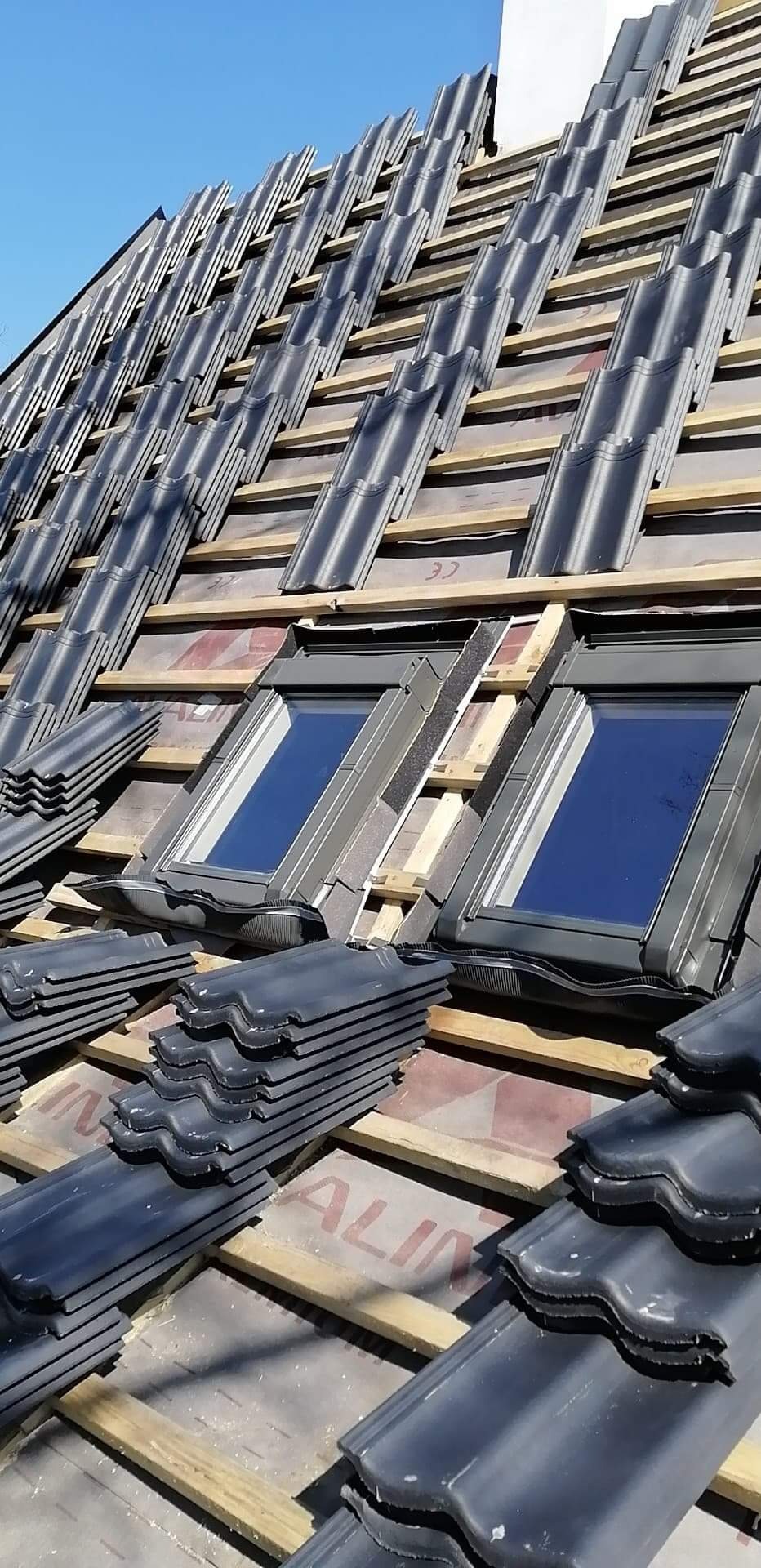 Montaż dachu z dachówką ceramiczną, widoczne okna dachowe i układ łat na folii izolacyjnej, w tle błękitne niebo.