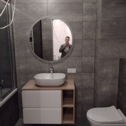 Tuszynski artur - Nowoczesna łazienka z szarymi płytkami, owalnym lustrem z podświetleniem LED, białą umywalką nablatową na szafce z półkami, i podwieszaną toaletą; odbicie osoby robiącej zdjęcie w lustrze.