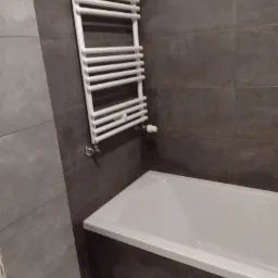 Nowoczesna łazienka z wanną obudowaną ciemnymi płytkami i białym grzejnikiem drabinkowym.