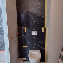 Tuszynski artur - Montaż wiszącej toalety na tle czarnej ściany z marmurowym wzorem, widoczne plastikowe krzyżyki dystansowe i taśma malarska zabezpieczająca krawędzie, ściana obok w trakcie szpachlowania.