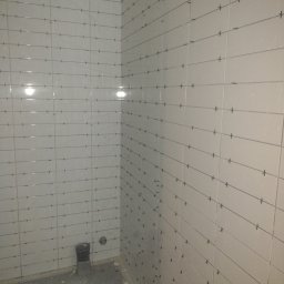 Tuszynski artur - Świeżo ułożone białe płytki na ścianach łazienki, z krzyżykami dystansowymi w spoinach, widoczny fragment podłogi w stanie surowym i odpływ kanalizacyjny.