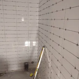 Świeżo ułożone, białe płytki na ścianach łazienki z krzyżykami dystansowymi, widoczny poziomica i wiadro z zaprawą, w tle rura odpływowa.