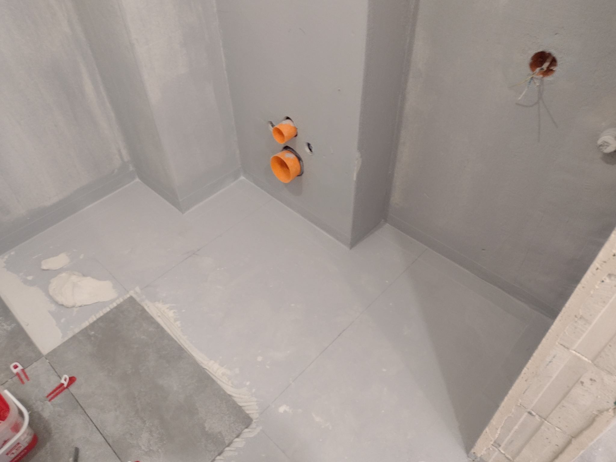 Przygotowana do położenia glazury łazienka w szarych odcieniach, widoczne fragmenty płytek, poziomowanie z użyciem klipsów, przygotowane otwory pod instalację hydrauliczną i elektryczną.