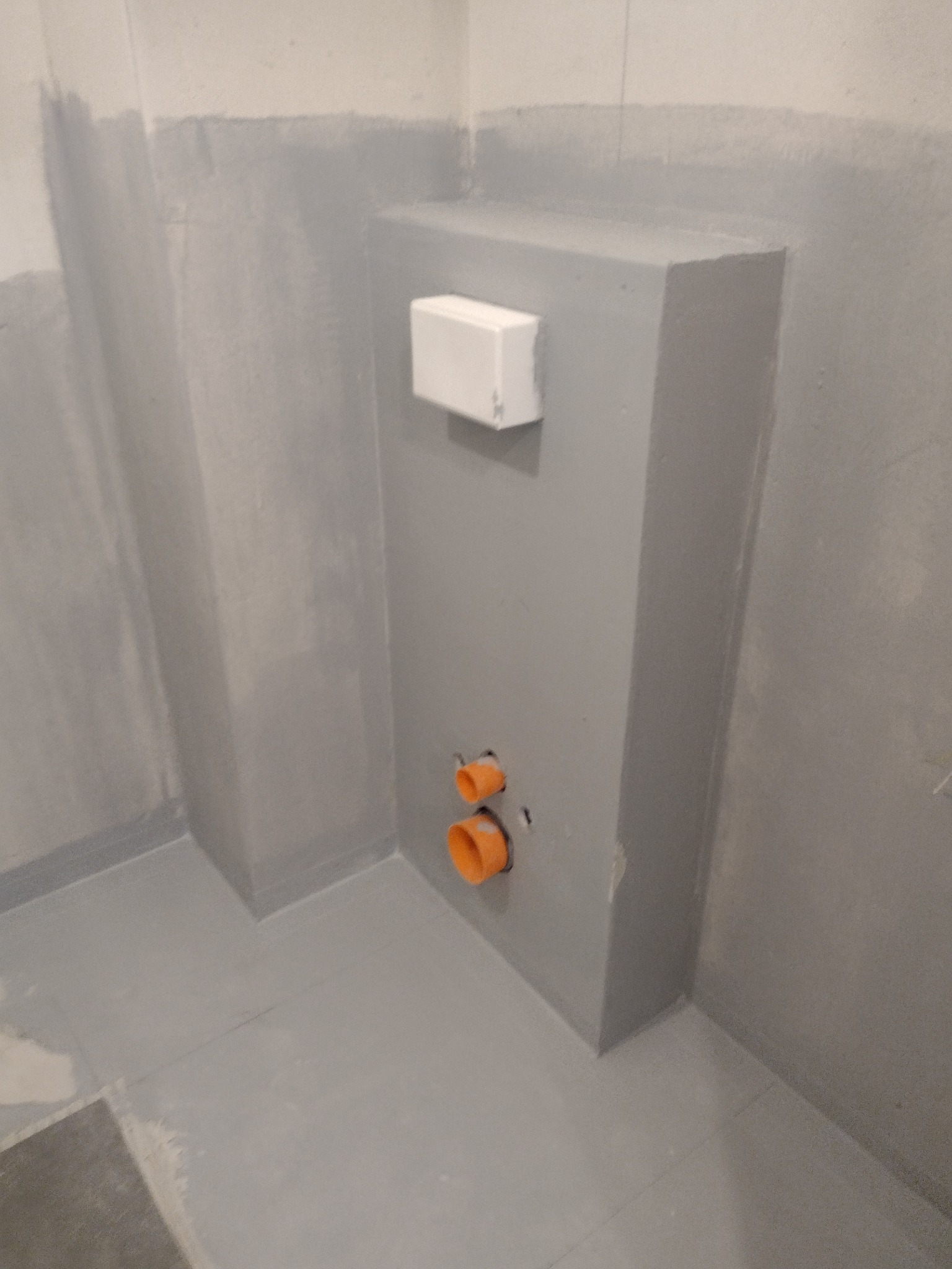 Szara ściana w łazience z zamontowanymi rurami kanalizacyjnymi i białą obudową stelaża podtynkowego WC, widoczne ślady po remoncie.