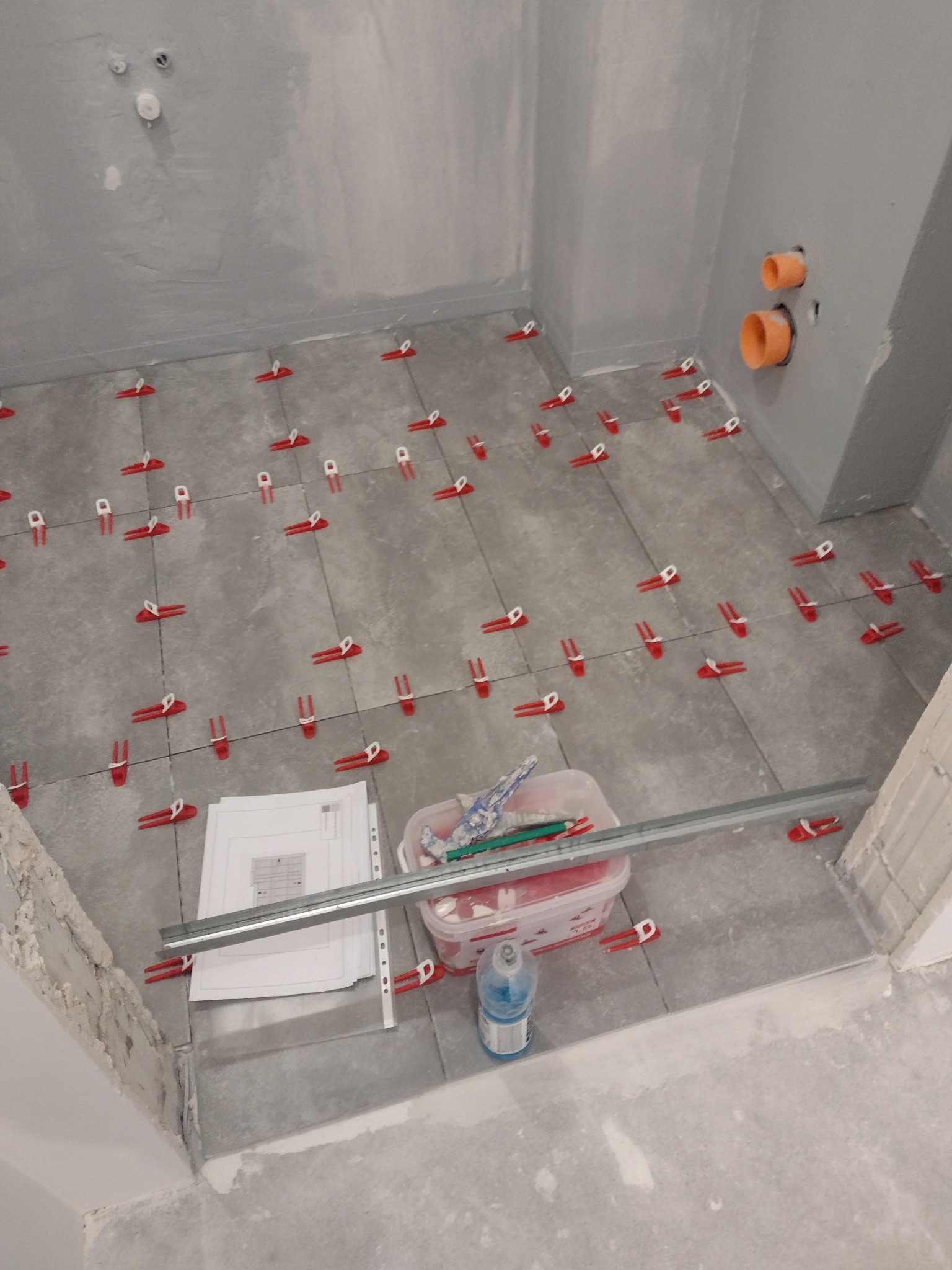 Szare płytki podłogowe w trakcie układania w łazience, widoczne czerwone klipsy poziomujące, narzędzia glazurnicze w pojemniku i plany na podłodze.