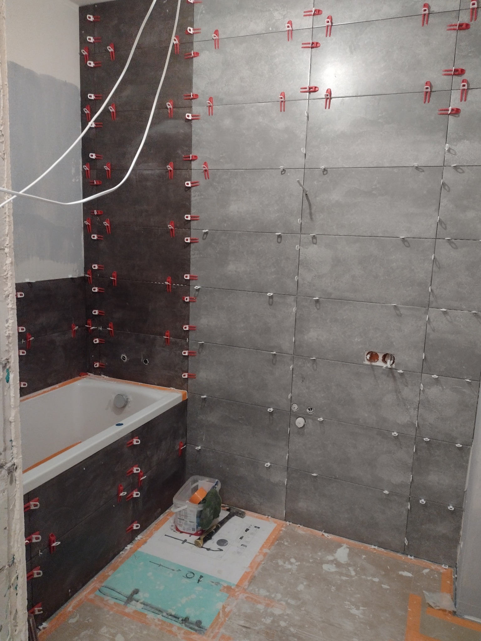 Montaż płytek w łazience, widoczne płytki w odcieniach szarości, czerwone klipsy poziomujące, przygotowana wanna, instalacja elektryczna, narzędzia i materiały na podłodze.