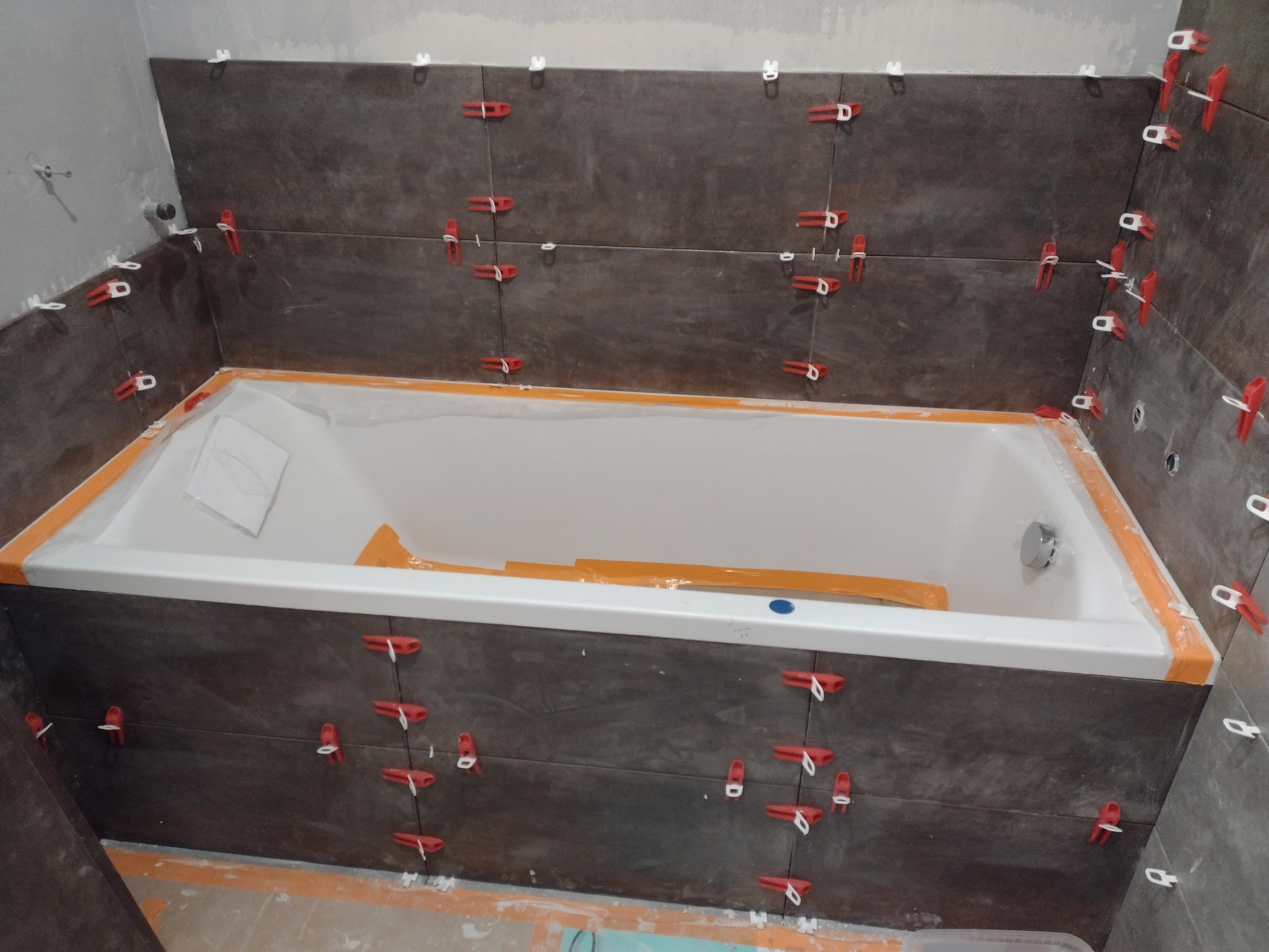 Montaż ciemnych, matowych płytek ceramicznych nad wanną, z użyciem czerwonych klinów poziomujących, na etapie układania.