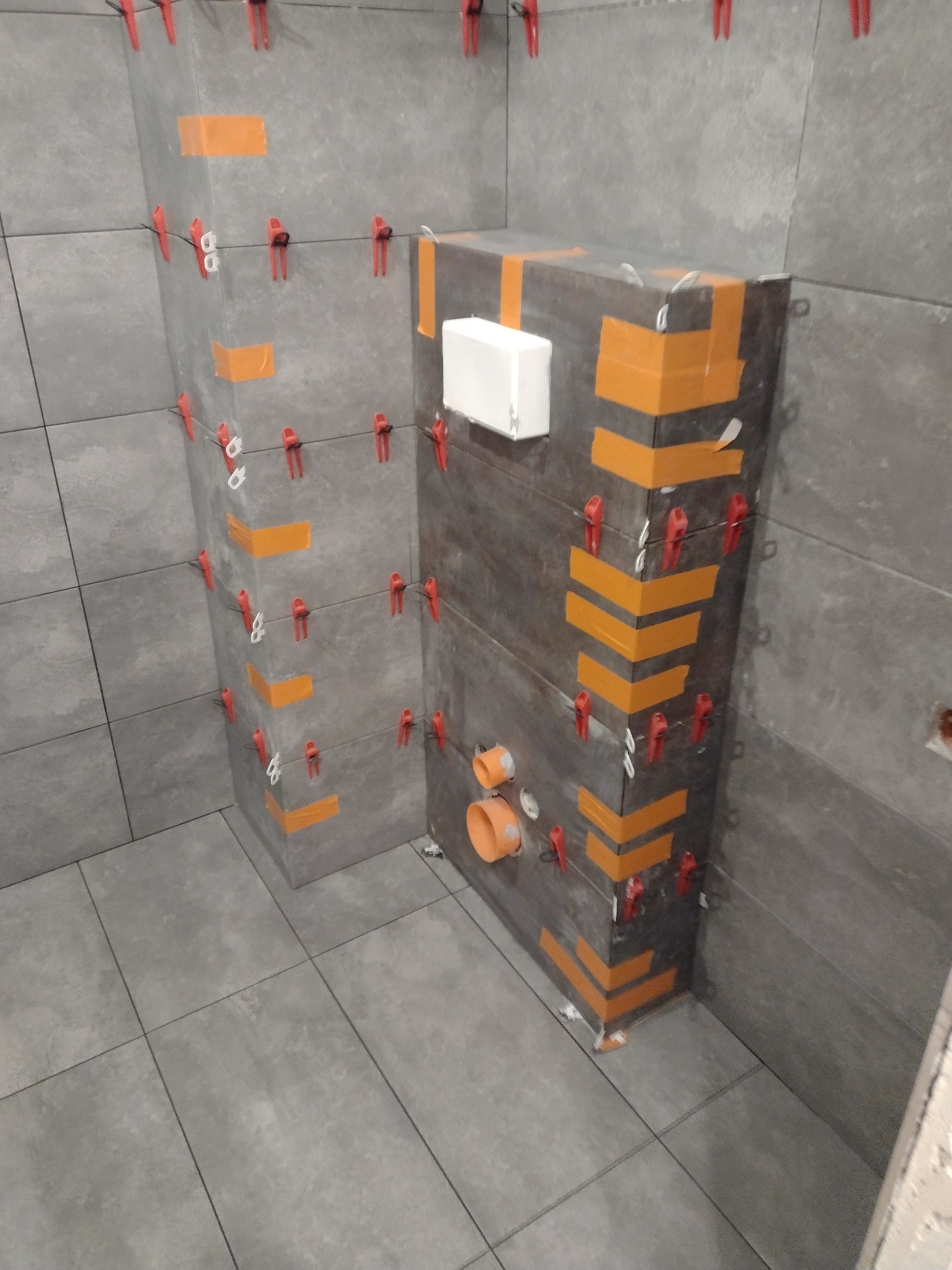 Szare płytki ceramiczne na ścianach i podłodze, częściowo zaklejone pomarańczową taśmą i zabezpieczone czerwonymi klipsami poziomującymi, widoczne rury kanalizacyjne.