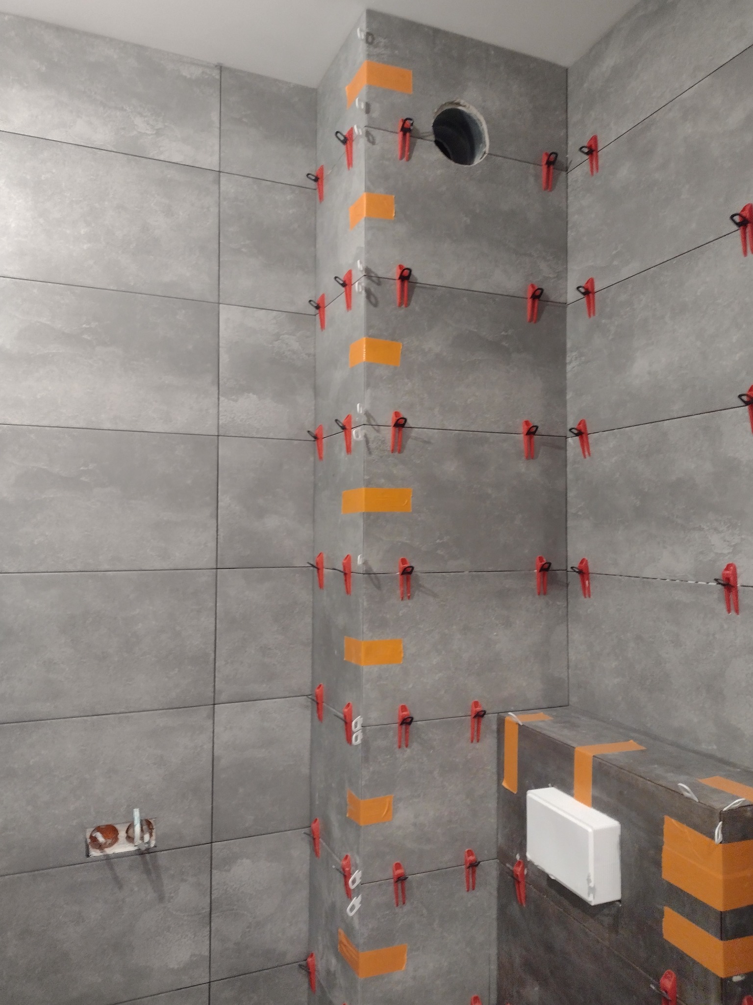 Świeżo ułożone, szare płytki w łazience, zabezpieczone pomarańczową taśmą i czerwonymi klipsami poziomującymi, widoczny otwór na rurę i przygotowane gniazdko elektryczne.