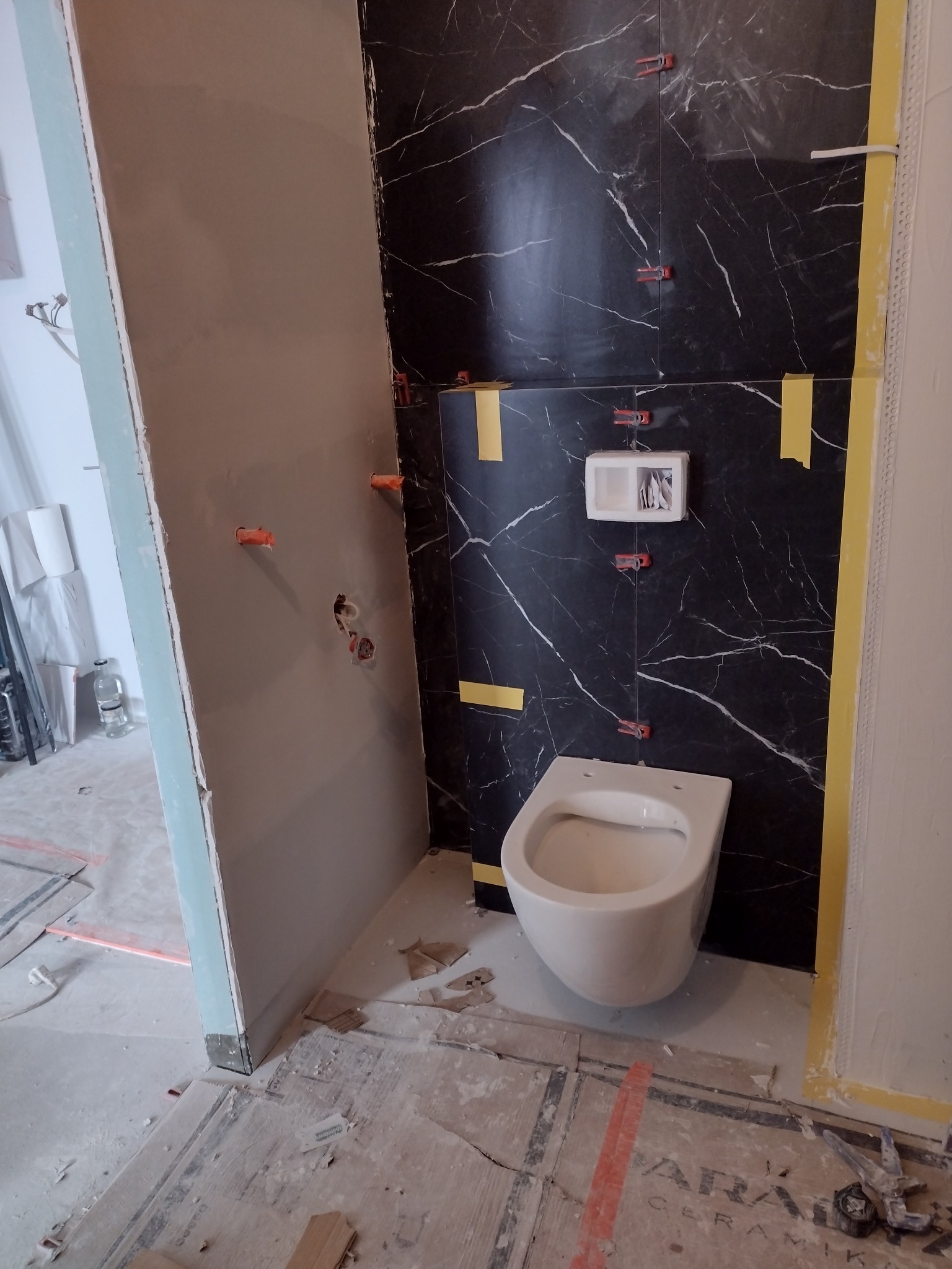 Wnętrze łazienki w trakcie remontu: montaż wiszącej toalety na tle czarnych płytek imitujących marmur, widoczne poziomice i elementy mocujące, surowy stan ścian z instalacją elektryczną.