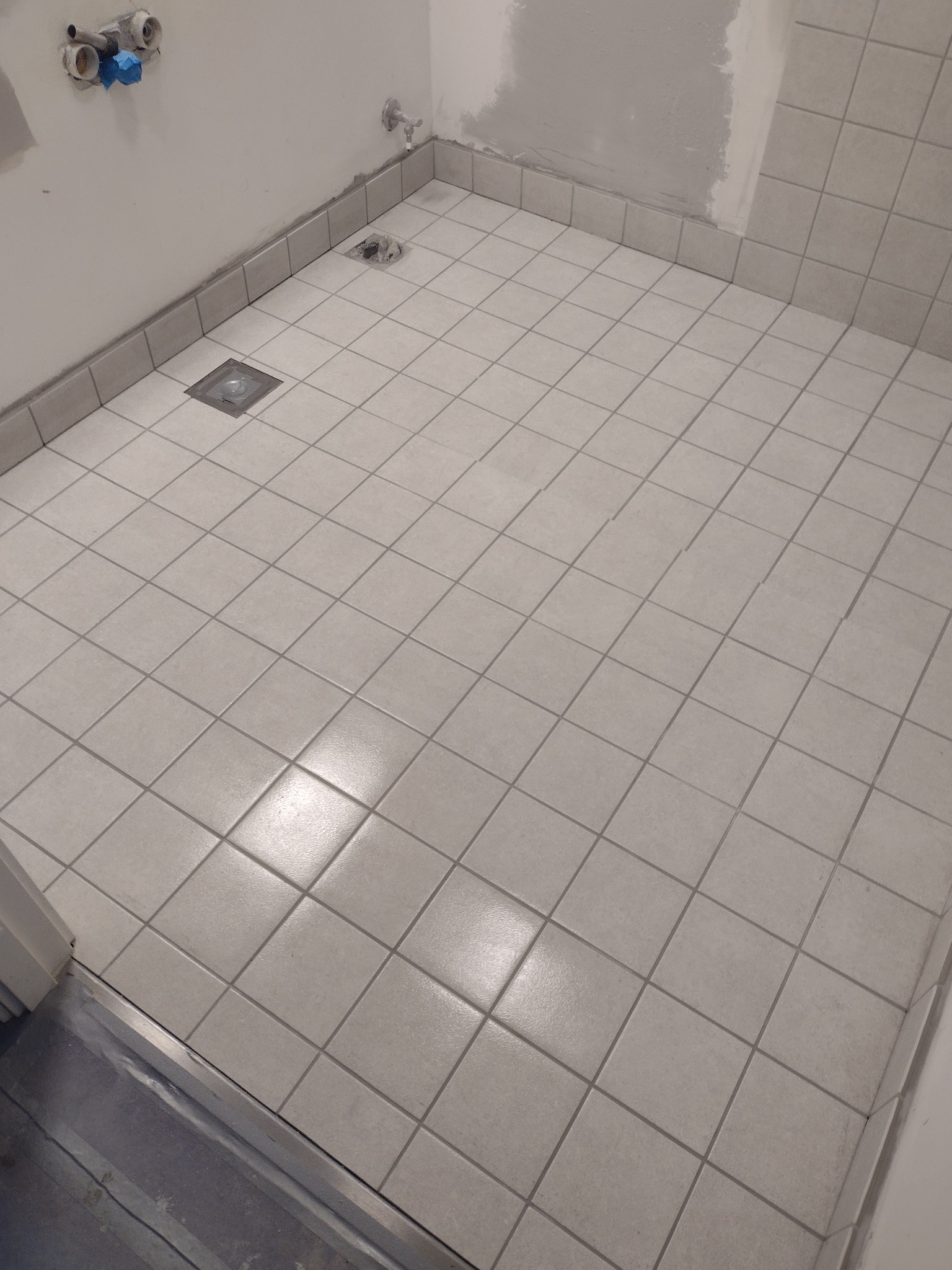 Ukończona podłoga w łazience wyłożona jasnymi, kwadratowymi płytkami ceramicznymi z widocznym odpływem liniowym i fragmentami ściany w stanie surowym.