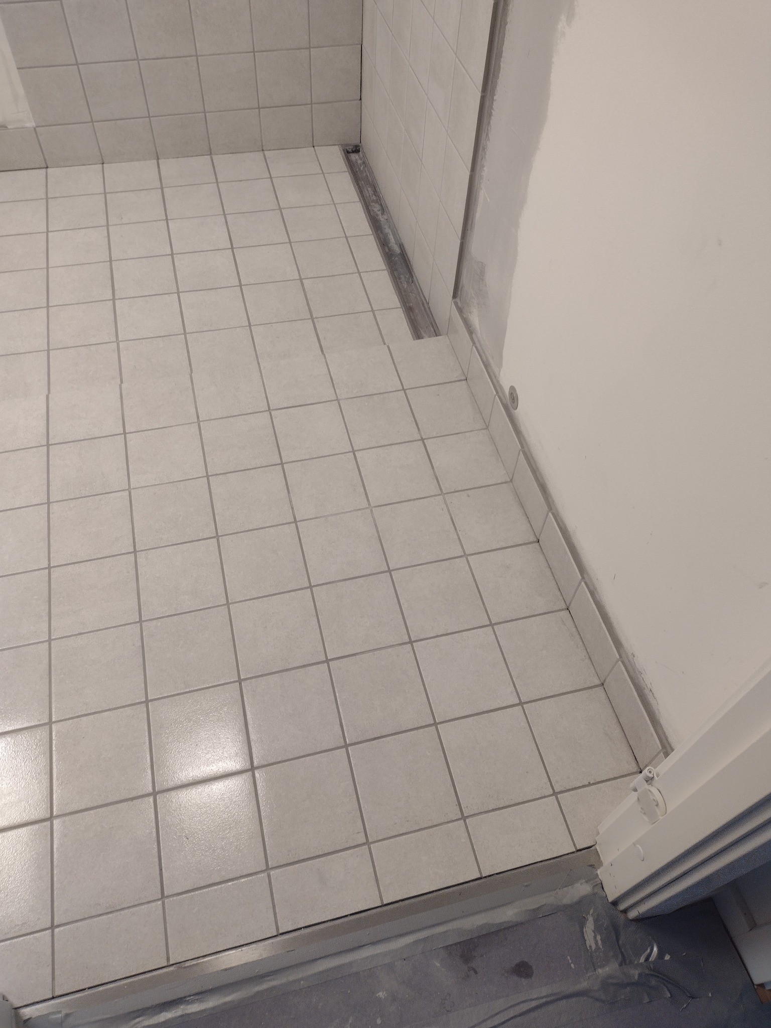 Jasne, kwadratowe płytki podłogowe w łazience, widoczny odpływ liniowy przy ścianie i częściowo wykończona ściana z widocznymi śladami szpachli.