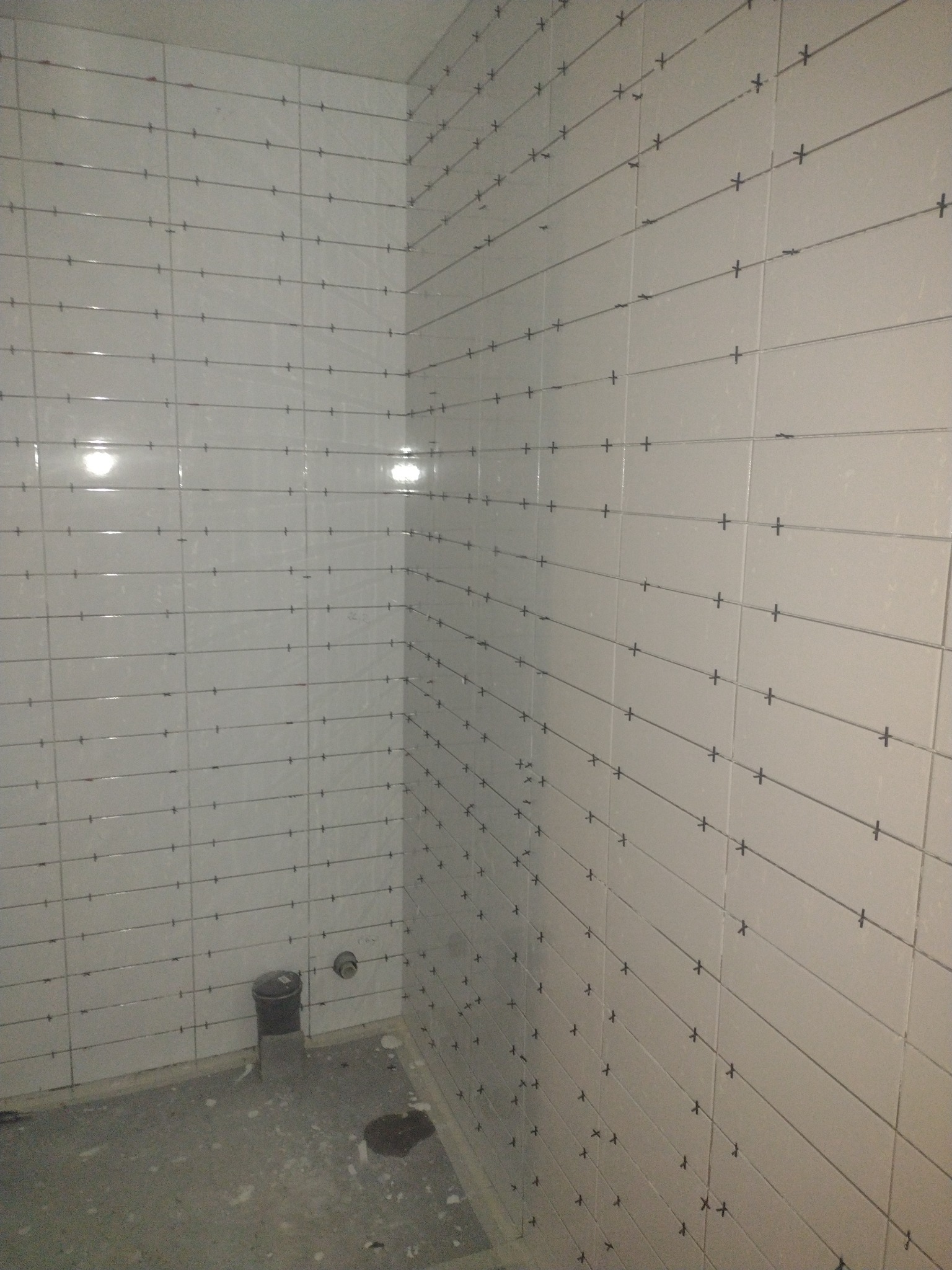 Świeżo ułożone białe płytki na ścianach łazienki, z krzyżykami dystansowymi w spoinach, widoczny fragment podłogi w stanie surowym i odpływ kanalizacyjny.