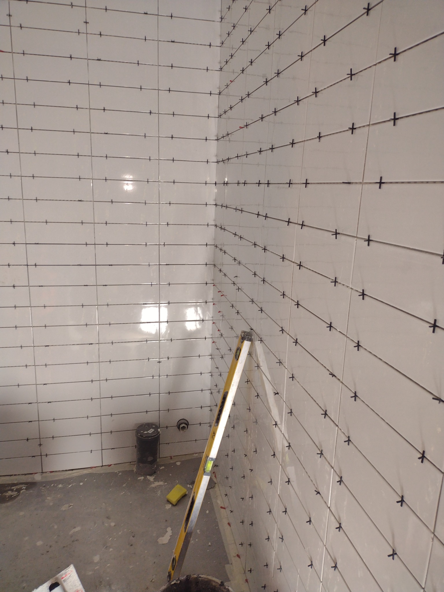 Świeżo ułożone, białe płytki na ścianach łazienki z krzyżykami dystansowymi, widoczny poziomica i wiadro z zaprawą, w tle rura odpływowa.